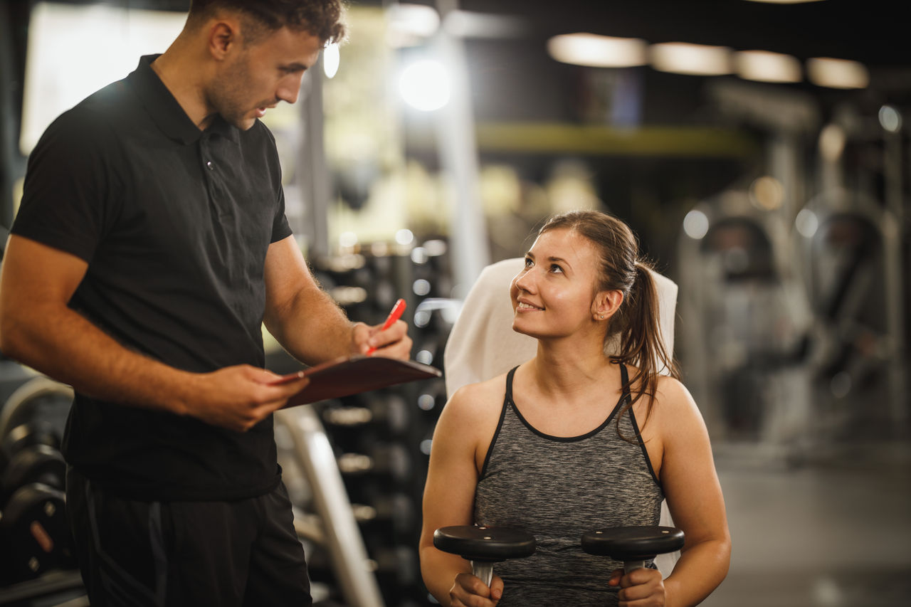 Shot of a muscular young woman in sportswear working out with personal trainer at the gym machine. She is pumping up her muscule with dumbbell.; Shutterstock ID 1837484953; purchase_order: POIIG0004097; job: OMG1193295; client: Danone UK; other: 480